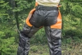 Nogawki ochronne Classic Husqvarna w kolorze czarnym z pomarańczowymi wstawkami, z widocznym regulowanym paskiem. Produkt prezentowany z tyłu, w pozycji stojącej. Otoczenie leśne, z drzewami w tle. Widziane detale bocznych kieszeni i logo producenta.