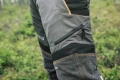 Spodnie ochronne do pasa Technical C Husqvarna w kolorze szaro-czarnym. Zbliżenie na łydkę, widoczny zamek błyskawiczny i elementy z materiału teksturowego. Wstawki z elastycznego materiału oraz logo na udzie. Tło z naturalną roślinnością.