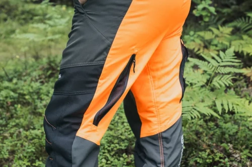 Spodnie ochronne Functional 20A Husqvarna w kolorze czarno-pomarańczowym, z zamkami błyskawicznymi po bokach. Ujęcie z boku, ukazujące detale konstrukcyjne i boczne kieszenie. Idealne do pracy w terenie, w naturalnym otoczeniu.