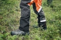 Spodnie ochronne Technical 20A Husqvarna w kolorze czarno-pomarańczowym, wykonane z wytrzymałego materiału. Ujęcie z boku, produkt leży na naturalnym podłożu. Widoczne odblaskowe detale na nogawkach oraz struktura materiału.