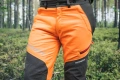 Spodnie ochronne Technical 20A Husqvarna w kolorze intensywnej pomarańczy z czarnymi elementami. Widoczny zamek boczny oraz logo Husqvarna na lewej nogawce. Produkt prezentowany w staniu na tle leśnym. Użyteczne do prac na świeżym powietrzu.