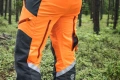 Spodnie ochronne Technical 20A Husqvarna, pomarańczowe z czarnymi wstawkami, odblaskowymi pasami na nogawkach, czarny zamek i kieszenie boczne. Widok z boku, detal i wykończenie w leśnym otoczeniu.