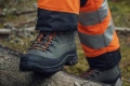Pomarańczowe spodnie ochronne Technical High Viz Husqvarna z odblaskowymi paskami, wytrzymały materiał. Widok z boku na osobę wspinającą się, detale szwów i sznurków. Model stoi na drewnianym pniu.