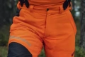 Spodnie ochronne Technical High Viz Husqvarna w intensywnej pomarańczy z czarnymi wstawkami, detal pasa z zapięciami i logo, gumowe mocowania na tle lasu podkreślającym ich użytkowy charakter.