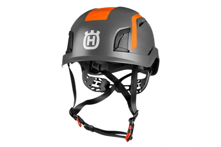 Kask dla arborystów Spire Vent Husqvarna w kolorze szarym z pomarańczowymi detalami. Ujęcie z boku, ukazujące zapięcie, wentylację oraz logo marki. Produkt przeznaczony do ochrony głowy podczas pracy na wysokości, z regulowanym paskiem.