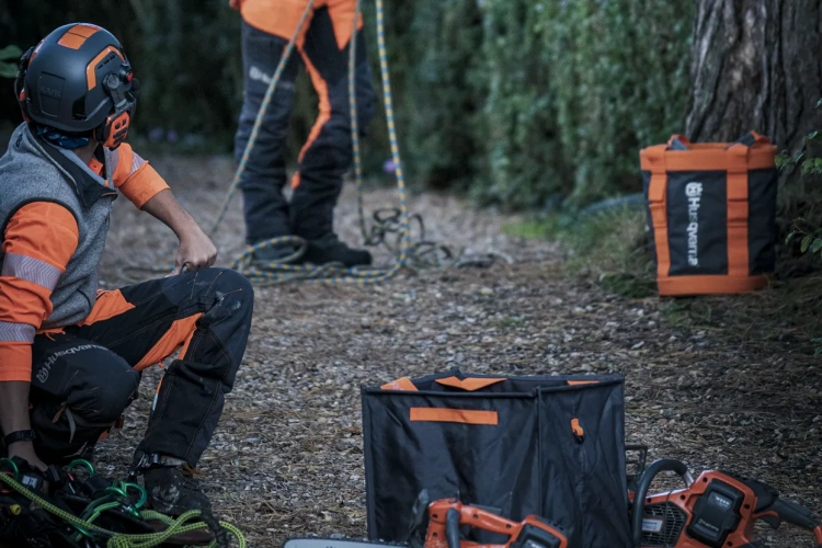 Kask dla arborystów Spire Vent Husqvarna, w kolorze ciemnoszarym z pomarańczowymi akcentami, widoczny z boku. Osoba w odzieży roboczej w pozycji klęczącej, w otoczeniu narzędzi oraz plecaka. W tle roślinność i fragment ścieżki.