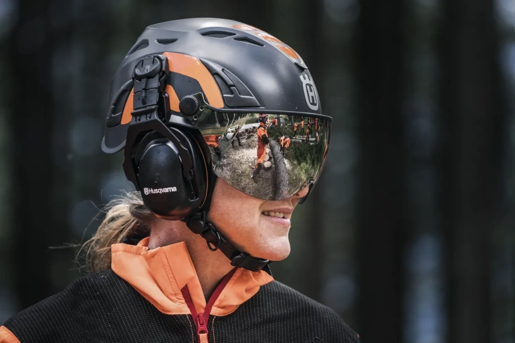 Kask dla arborystów Spire Vent Husqvarna w kolorze czarnym z pomarańczowymi akcentami, z zamontowanymi ochronnymi nausznikami. Ujęcie frontalne, na twarzy użytkownika z refleksją na wizjerze. Dobrze widoczne logo Husqvarna, kask w terenie leśnym.