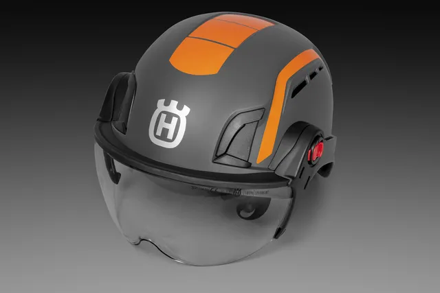 Kask arborystyczny Spire Vent Husqvarna w kolorze szarym z pomarańczowymi akcentami, z osłoną twarzy. Widok z góry, stojący na ciemnym tle. Wygodne mocowanie i wentylacja, logo Husqvarna widoczne z przodu.