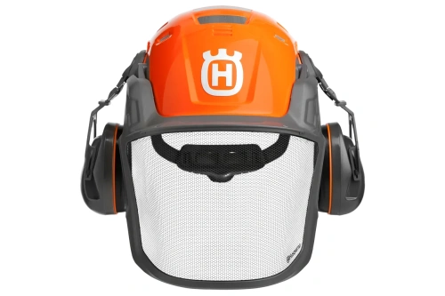 Kask leśny Husqvarna w kolorze pomarańczowym i szarym, z siatką ochronną z przodu i słuchawkami po bokach. Ujęcie frontalne ukazuje logo Husqvarna na górze. Kask jest zamocowany na płaskiej powierzchni.