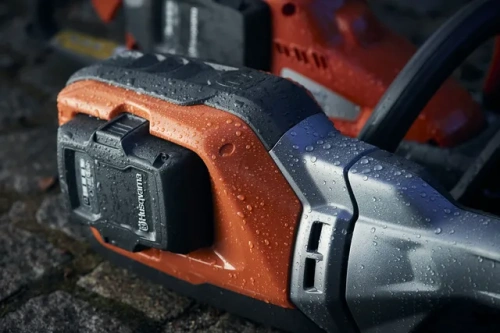 Podkrzesywarka akumulatorowa Husqvarna 530iPT5 w zbliżeniu, pokazująca pomarańczową obudowę z szarymi elementami i przyłączonym akumulatorem. Widoczne kropelki wody na powierzchni, co sugeruje warunki atmosferyczne przy fotografowaniu.