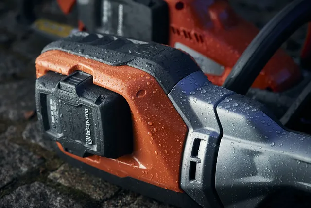 Podkrzesywarka akumulatorowa Husqvarna 530iPT5 w zbliżeniu, pokazująca pomarańczową obudowę z szarymi elementami i przyłączonym akumulatorem. Widoczne kropelki wody na powierzchni, co sugeruje warunki atmosferyczne przy fotografowaniu.