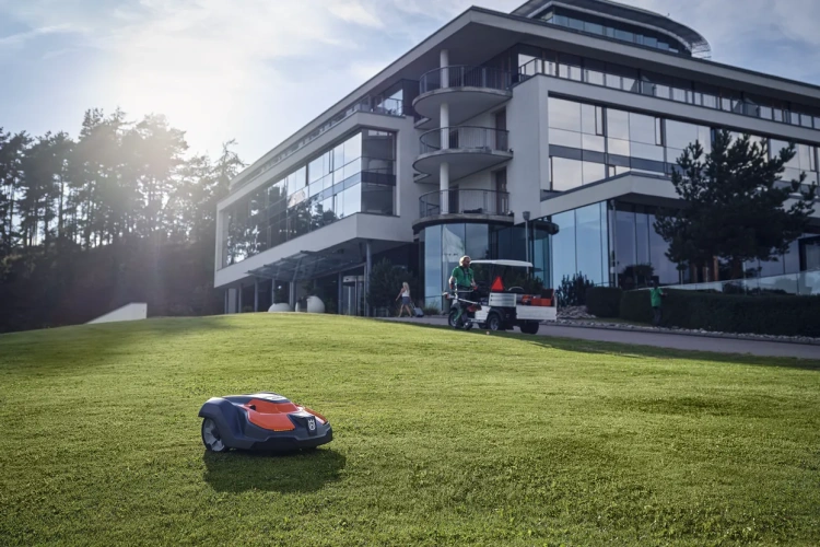 Robot koszący Automower® 550 Epos w kolorze szarym i pomarańczowym, ukazany z bocznego ujęcia na zielonej trawie. W tle nowoczesny budynek z dużymi oknami. Widać również osobę obsługującą pojazd do transportu.