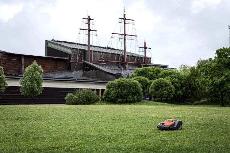 Robot koszący Automower® 550 Epos Husqvarna w pozycji leżącej na zielonej trawie, z widocznym pomarańczowym deklem i szarym korpusem. Ujęcie z boku, w tle widoczna architektura budynku z masztami. Teren jasno oświetlony, z drzewami w otoczeniu.