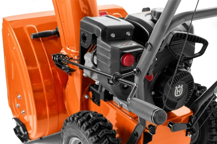 Pomarańczowa odśnieżarka ST124 Husqvarna, widok z boku. Metalowa obudowa, regulowany uchwyt z gumą, czerwony przycisk, silnik i duże bieżnikowane koła na białym tle podkreślającym detale.