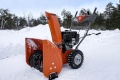 Pomarańczowa odśnieżarka ST124 Husqvarna na zaśnieżonym terenie, z widocznym wirnikiem, gumowymi oponami z bieżnikiem, uchwytem z przyciskami i logo Husqvarna, na tle zaśnieżonego lasu.