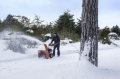 Odśnieżarka ST124 Husqvarna w akcji, operator stoi obok, skierowana w lewo. Wyrzuca śnieg w powietrze. Tło to zalesiony teren pokryty śniegiem, z około drzewami. Operator ubrany w ciemną odzież, akcesoria na odśnieżarce są widoczne.
