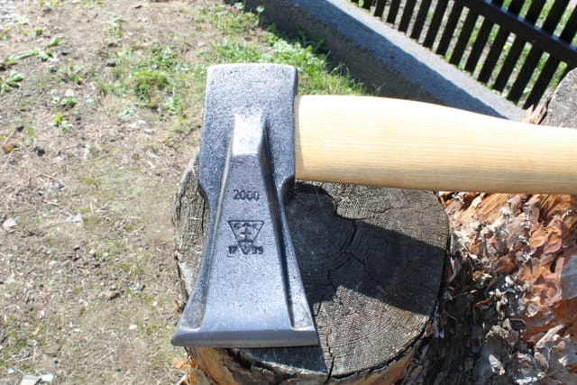 Siekiera klinowa 2 kg Krumpholz z drewnianą rączką leży na pniu. Metalowa, matowa głowica z oznaczeniem 2000 i logo. Ujęcie z boku ukazuje ostrze. Tło naturalne z drobną roślinnością.