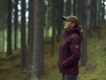 Damska wodoodporna kurtka Husqvarna Xplorer w kolorze burgundowym z kapturem, noszona z boku przez kobietę w lesie, z logo po lewej stronie. Tło: drzewa i zieleń podkreślające outdoorowy charakter kurtki.