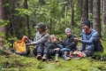 Rodzina siedzi na zielonym mchu w lesie. Dorośli mają na sobie odzież outdoorową w odcieniach szarości i niebieskiego, dzieci w swetrach z kapturem. Wszyscy mają akcesoria turystyczne, a obok widoczne są plecaki oraz termosy. Atmosfera relaksu.