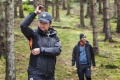 Kurtka damska Husqvarna Xplorer Shell w kolorze czarnym z pomarańczowymi detalami, noszona przez kobietę w lesie. Ujęcie z boku, zbliżenie na rękę regulującą rękaw. Tło z drzewami i zieloną trawą, wzmacnia kontekst aktywności outdoorowej.