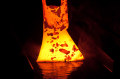 Zbliżenie na gorące ostrze siekieromłotu w czarnym tle, z jasno-pomarańczowym i czerwonym cieniowaniem. Widoczne detale ostrza i nieregularne krawędzie, z parą w tle sugerującą proces formowania.