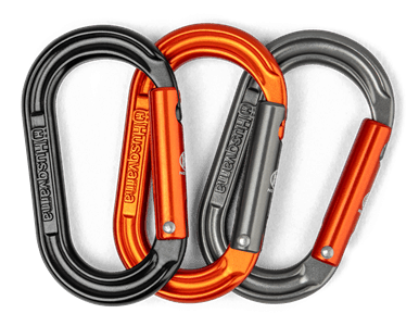 Zestaw trzech karabińczyków Husqvarna w kolorach: czarnym, szarym i pomarańczowym. Wszystkie mają ergonomiczną formę i stalowe zapięcia. Ułożone obok siebie, ukazują różne odcienie metalu oraz logo Husqvarna na każdym z karabińczyków.