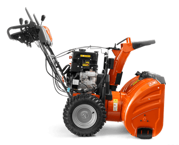 Odśnieżarka ST330 Husqvarna w pomarańczowym kolorze, z silnikiem widocznym z boku. Produkt stoi w pozycji pionowej, z metalowym, teksturowanym zbiornikiem. Wyposażona w gumowe koła oraz uchwyt do sterowania. Tło neutralne, jednolite.