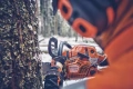 Pilarka Husqvarna 562 XP® Mark II w pomarańczowym kolorze, z metalowym ostrzem, trzymana przez użytkownika w rękawicach. Fotografia z boku, pokazująca detale obudowy i logo producenta, w tle widoczny pień drzewa pokryty mchem, w zimowym otoczeniu.