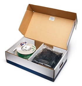 Zestaw instalacyjny Automower® w otwartym kartonie. Wewnątrz widoczne są: zielona linka obwodowa, zapasowe złącza oraz czarne haki montażowe. Karton ma prostą, białą strukturę z niebieskimi detalami na spodzie.