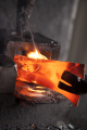Widok na gorący metal w piecu, intensywnie świecący pomarańczowym kolorem. Metalowa forma trzymana przez narzędzie, umieszczona nad płomieniami. W tle czarna struktura pieca oraz unoszący się dym.