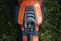 Przycisk sterujący pilarki akumulatorowej Husqvarna 542i XP, w kolorze pomarańczowym, z widocznym panelem wyświetlacza. Widać ikony stanu baterii, ostrzeżenie oraz przyciski zasilania. Kąt ujęcia z góry, na tle zielonej trawy.