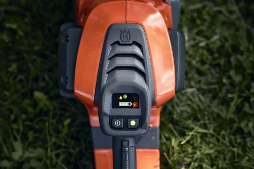 Przycisk sterujący pilarki akumulatorowej Husqvarna 542i XP, w kolorze pomarańczowym, z widocznym panelem wyświetlacza. Widać ikony stanu baterii, ostrzeżenie oraz przyciski zasilania. Kąt ujęcia z góry, na tle zielonej trawy.