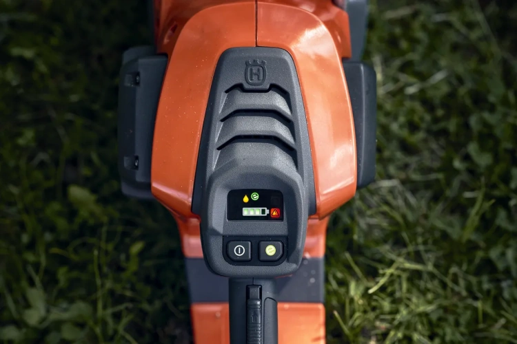 Przycisk sterujący pilarki akumulatorowej Husqvarna 542i XP, w kolorze pomarańczowym, z widocznym panelem wyświetlacza. Widać ikony stanu baterii, ostrzeżenie oraz przyciski zasilania. Kąt ujęcia z góry, na tle zielonej trawy.