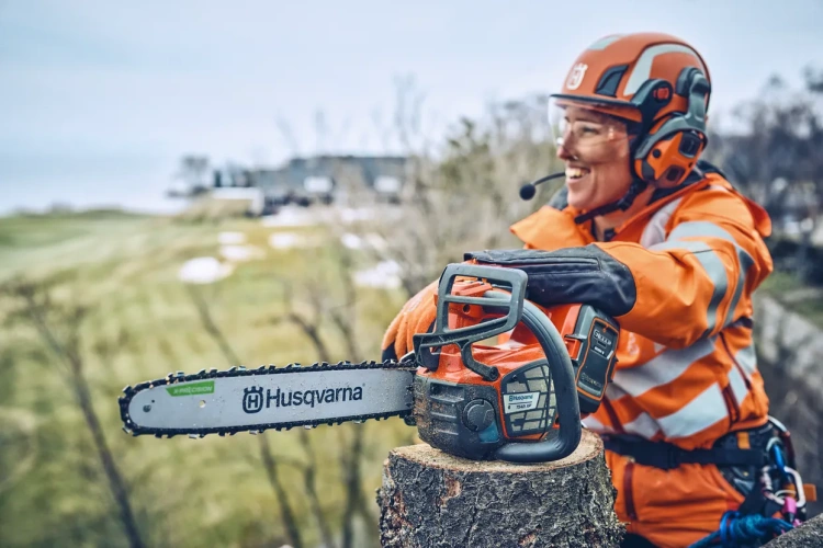 Pomarańczowa pilarka akumulatorowa Husqvarna T542i XP na ściętym pniu drzewa, widok z boku z detalami łańcucha i logo, z osobą w odzieży roboczej, na tle drzew i trawnika.