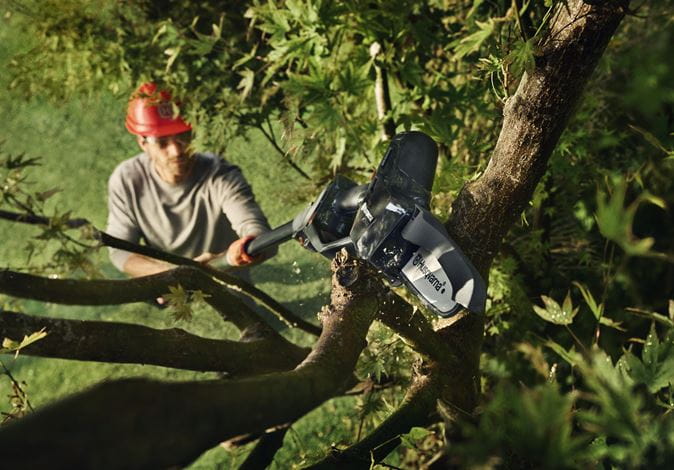 Podkrzesywarka Husqvarna Aspire™ PE5-P4A 2P w użyciu, skierowana na gałęzie drzewa. Użytkownik w szarej koszulce i kasku ochronnym trzyma urządzenie. Tło przedstawia zieleń liści i gałęzi, podkreślając kontekst pracy w ogrodzie.