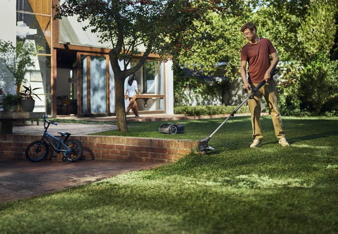 Podkaszarka Aspire™ T28-P4A 1P Husqvarna w rękach użytkownika, stojącego na zadbanym trawniku. W tle widoczny stół z ogrodowymi meblami i zdjęcie roweru. Produkt w kolorze czarnym z metalowymi akcentami, wykonany z tworzywa sztucznego.