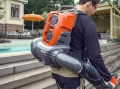 Dmuchawa akumulatorowa Husqvarna 340iBT na plecach użytkownika, pomarańczowa z szarymi elementami i elastyczną rurą. Marka Husqvarna widoczna na obudowie. Tło jasne, basen i parasol.