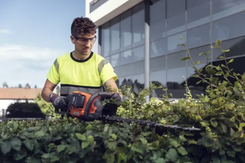 Nożyce akumulatorowe Husqvarna 522iHD60 w użyciu. Użytkownik w żółtej koszulce z ciemnym paskiem przycina żywopłot. W tle szklany budynek. Widać detale narzędzia: pomarańczowy korpus, metalowe ostrza i ergonomiczny uchwyt.
