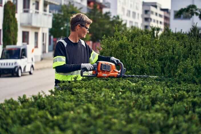 Mężczyzna w odzieży roboczej z odblaskami używa pomarańczowo-czarnych nożyc akumulatorowych Husqvarna 522iHDR60 do przycinania gęstej zieleni na tle miejskiej scenerii.
