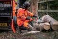 Pilarka akumulatorowa Husqvarna 540i XPG w użyciu, operator w pomarańczowym kombinezonie ochronnym, ścina pnie drzew na ziemi, tło z drewnianym ogrodzeniem i przyczepą. Widoczne detale piły oraz zastosowanie w terenie leśnym.