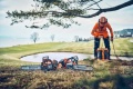 Pilarka akumulatorowa Husqvarna 542i XPG leży na zielonej trawie, obok stoi operator ubrany w pomarańczowy kombinezon ochronny. Tło przedstawia staw i zalesiony teren. Na pilarkach widoczne logo Husqvarna oraz detale konstrukcyjne.