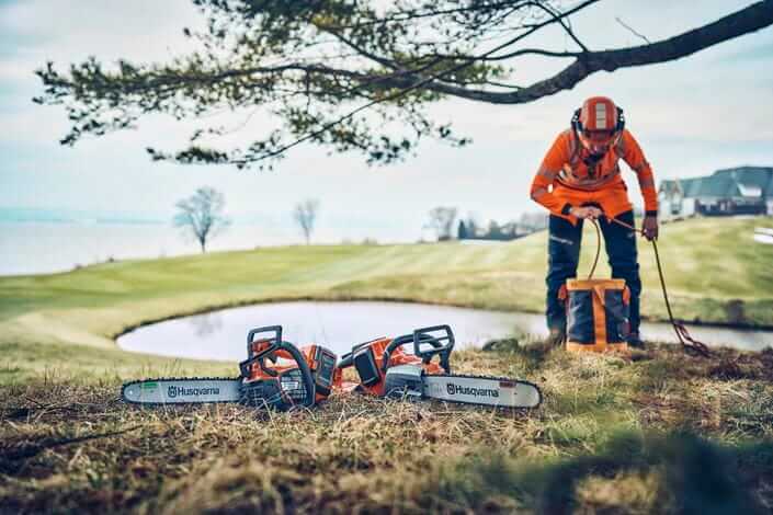 Pilarka akumulatorowa Husqvarna 542i XPG leży na zielonej trawie, obok stoi operator ubrany w pomarańczowy kombinezon ochronny. Tło przedstawia staw i zalesiony teren. Na pilarkach widoczne logo Husqvarna oraz detale konstrukcyjne.