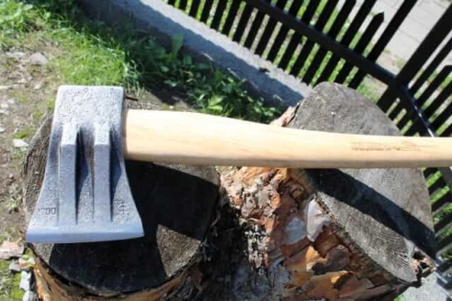 Siekiera 2-klinowa 1,7 kg Krumpholz na tle ciemnego kłody drewna. Kąt ujęcia z góry, z bliska, pokazuje stalowe ostrze z wyraźnymi klinami oraz drewniany trzon w naturalnym kolorze. Tło z zieloną trawą i drewnianą siatką.