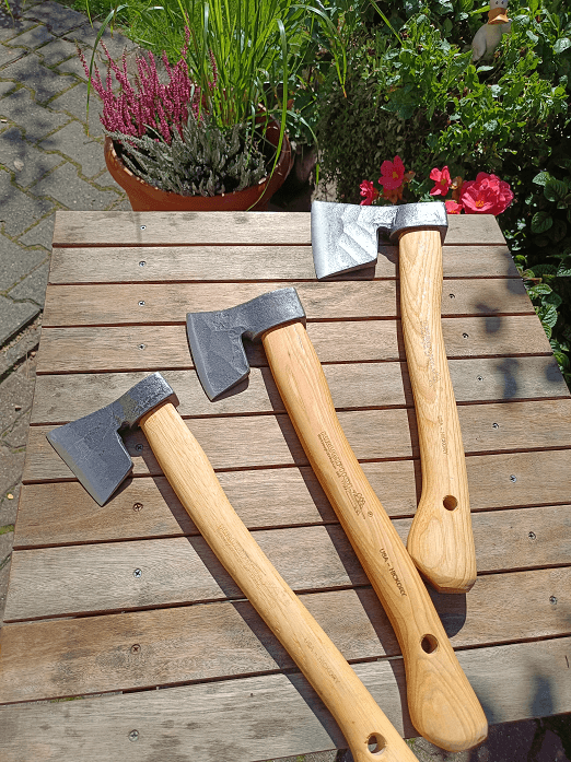 Toporek leśny Krumpholz o długości 38 cm, wykonany z metalu i drewna, trzy egzemplarze na drewnianym blacie. Ujęcie z góry, ukazujące metalowe ostrze i drewniane trzonki. Tło z zielenią roślin oraz kwiatami.