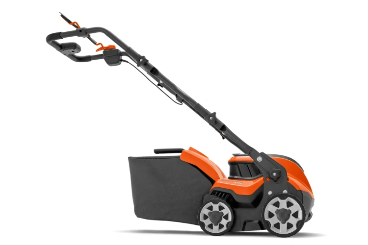 Wertykulator akumulatorowy S138i Husqvarna, boczny widok. Ergonomiczna rączka, pomarańczowy korpus, czarne detale, kosz na trawę, stabilne koła z wzorem na jednolitym tle.