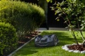 Robot koszący Husqvarna Automower® 310E NERA w kolorze czarnym, widoczny na trawie. Ujęcie z boku, w terenie ogrodowym, z krzewami i dekoracyjny kamiennym obrzeżem. Produkt leży na trawie, bez dodatkowych elementów montażowych.