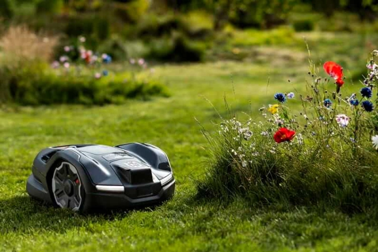 Robot koszący Husqvarna Automower® 410XE NERA w kolorze czarnym, z okrągłym korpusem i dużymi kołami, prezentowany w ogrodzie. Ujęcie z boku pokazuje go w ruchu, na tle zielonej trawy i kolorowych kwiatów.