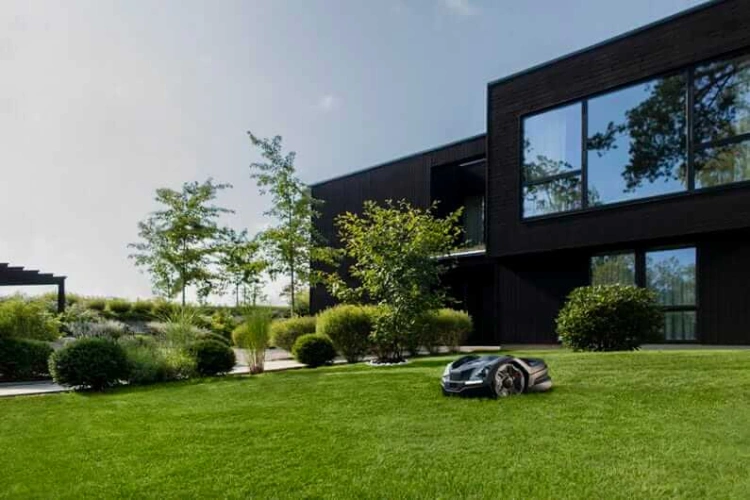 Robot koszący Husqvarna Automower® 410XE NERA w ogrodzie, na zielonym trawnikiem. Ujęcie z boku, pokazujące detale obudowy w kolorze antracytowym oraz koła. W tle nowoczesny dom oraz roślinność, na prostym, jasnym tle nieba.