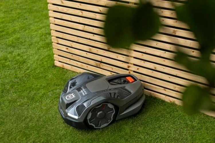 Robot koszący Husqvarna Automower® 410XE NERA w kolorze szarym, z widocznymi detalami, takimi jak logo Husqvarna i przycisk startowy w pomarańczowym kolorze. Produkt leży na trawie, z widokiem na boczną część, w tle drewniana struktura.