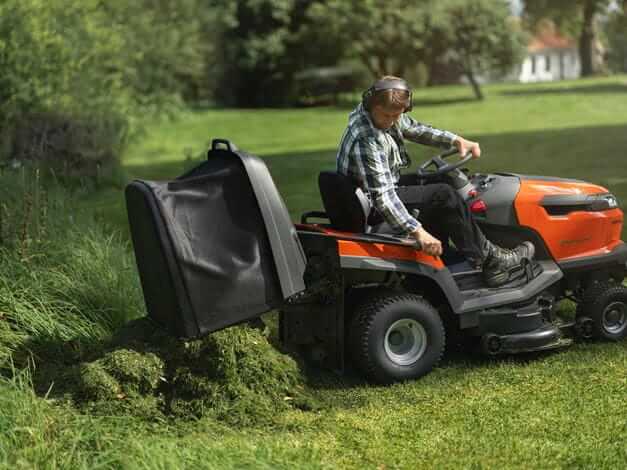 Traktor TC 220T Husqvarna w kolorze pomarańczowym z czarnym siedzeniem, widoczny w akcji na trawniku. Operator w koszuli w kratę wyrzuca skoszoną trawę z tylnego zbiornika. Ujęcie z boku, ukazujące detal pracy urządzenia w otoczeniu zieleni.